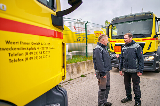 Zwei Weert Ihnen Mitarbeiter stehen vor einem gelben Firmen-LKW und unterhalten sich freundlich.